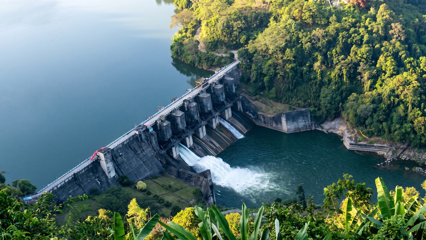 Aerial view of the Ranganadi Dam releasing water through open spillway gates into the river below, with its reservoir and forested hills around it