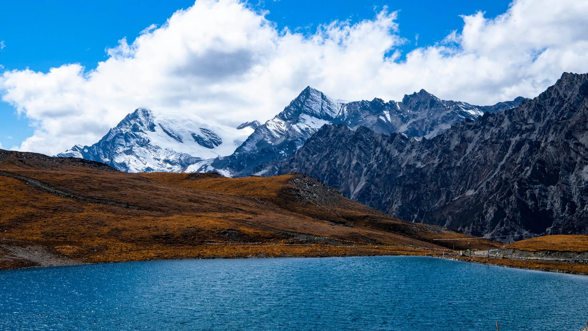 A blue mountain lake beneath brown alpine slopes, snow-covered peaks and clouds